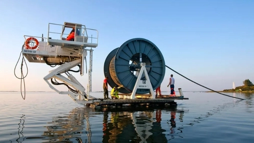 Crew working on a cable-laying barge with a large drum and scissor lift platform on calm coastal water