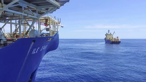 Large blue marine vessel in the foreground with a support ship visible in the distance on calm open water