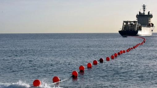 Cable-laying vessel at sea trailing a line of red buoys marking a subsea cable route across calm open water