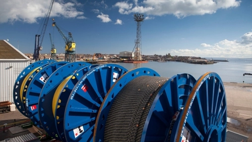 Large blue cable reels on a port quayside with harbour cranes and a coastal town visible in the background