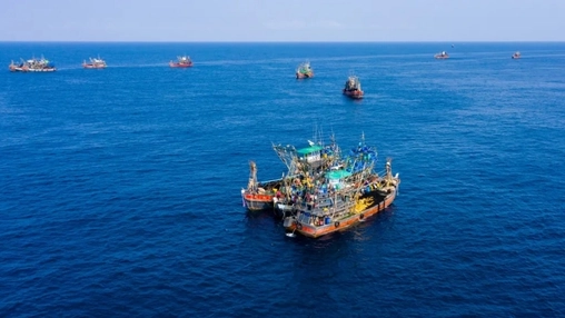Fishing trawlers clustered on blue ocean seen from above, with additional vessels spread across the horizon