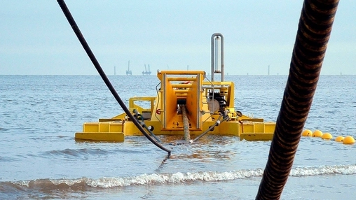 Yellow cable-laying machine in shallow coastal waters, with an offshore industrial platform on the horizon